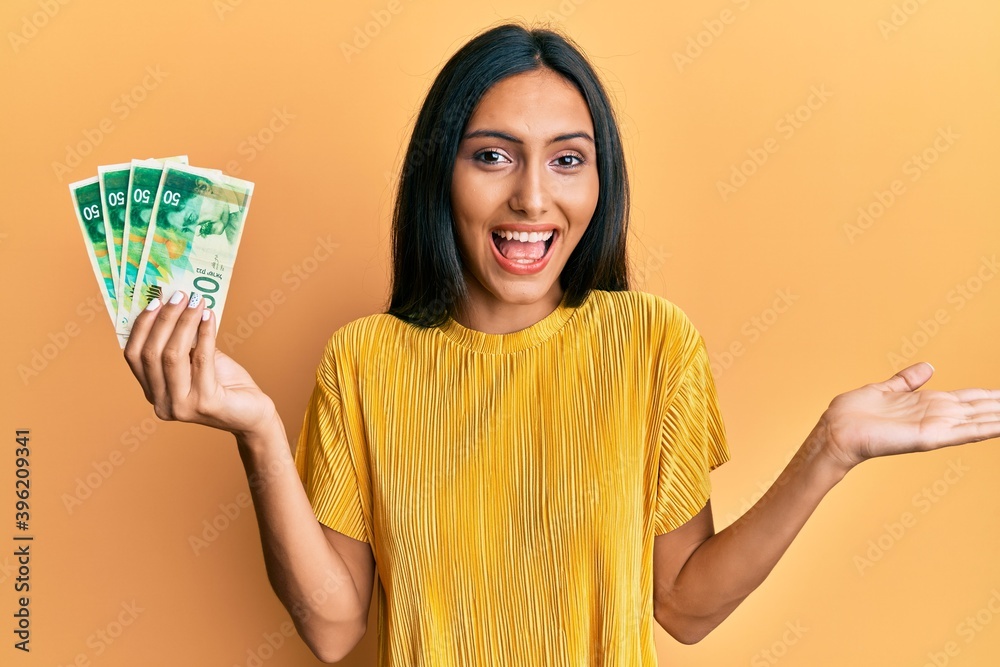 Young brunette woman holding 50 israel shekels banknotes celebrating achievement with happy smile and winner expression with raised hand Young brunette woman holding 50 israel shekels banknotes celebrating achievement with happy smile and winner expression with raised hand