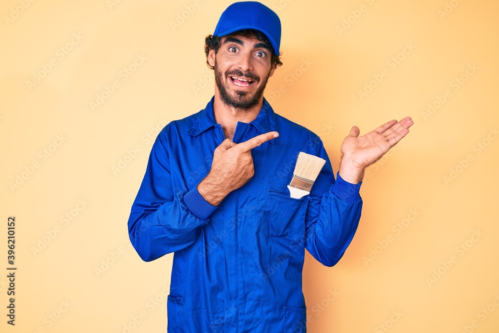 Handsome young man with curly hair and bear wearing builder jumpsuit uniform amazed and smiling to the camera while presenting with hand and pointing with finger.