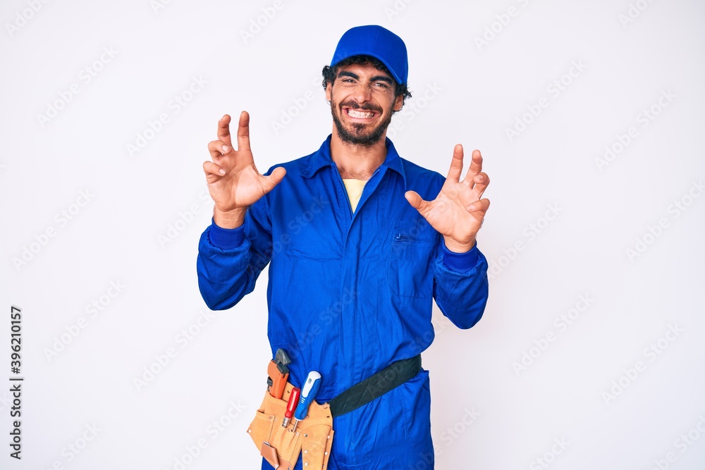 Handsome young man with curly hair and bear weaing handyman uniform smiling funny doing claw gesture as cat, aggressive and sexy expression