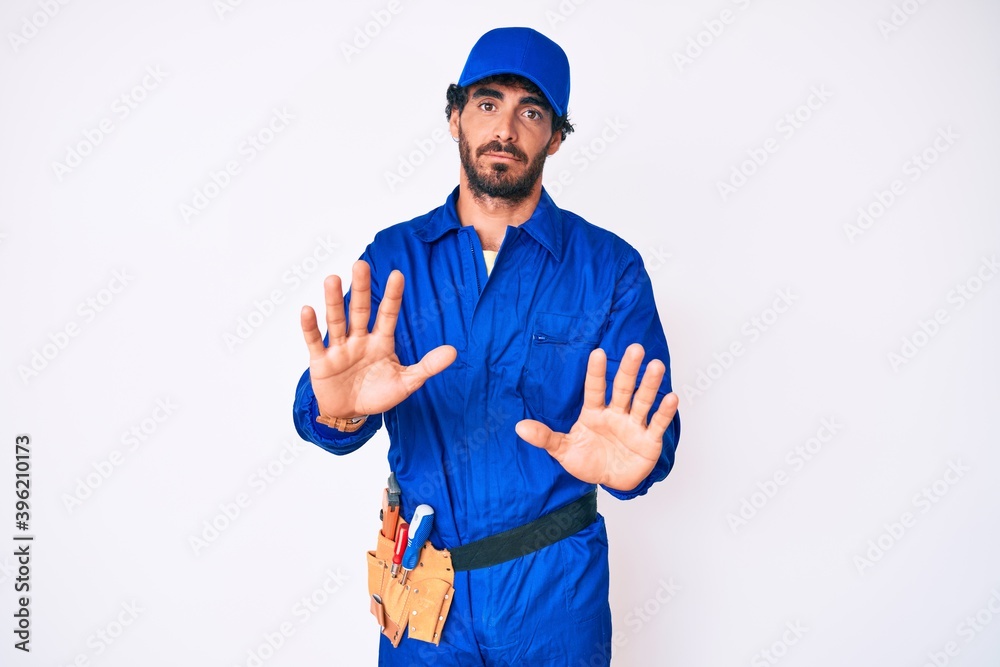 Handsome young man with curly hair and bear weaing handyman uniform moving away hands palms showing refusal and denial with afraid and disgusting expression. stop and forbidden.