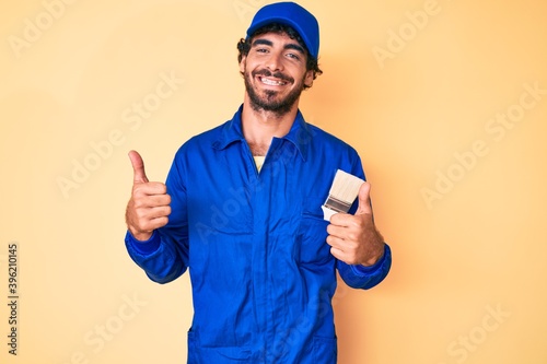 Handsome young man with curly hair and bear wearing builder jumpsuit uniform success sign doing positive gesture with hand, thumbs up smiling and happy. cheerful expression and winner gesture.