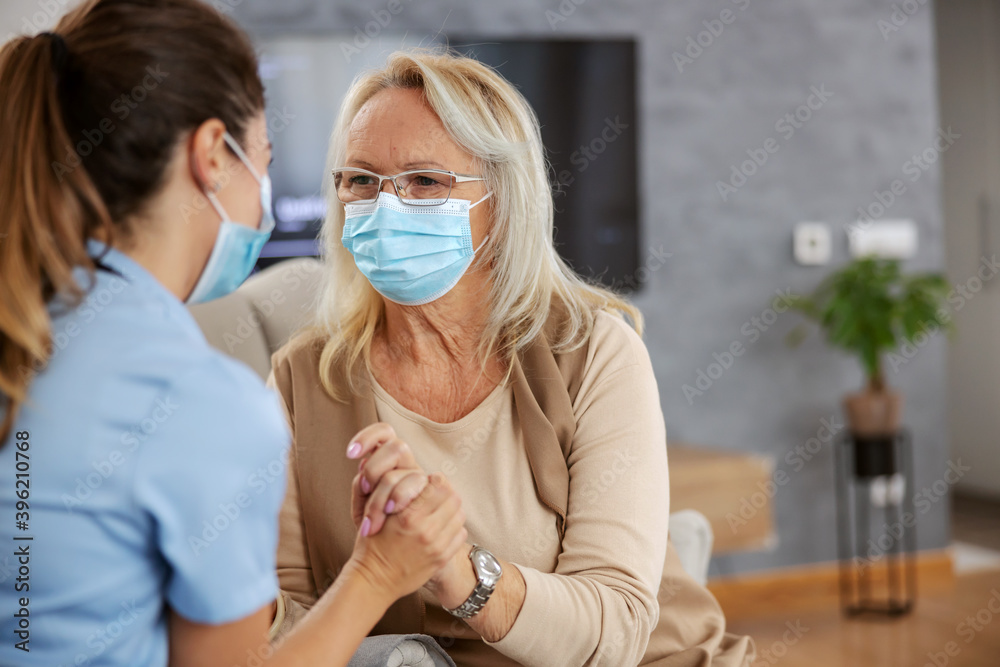 Senior woman with face mask sitting at home and listening advices from nurse during corona virus outbreak.