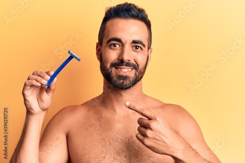 Young hispanic man standing shirtless holding razor smiling happy pointing with hand and finger