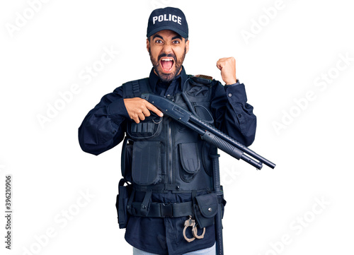 Young hispanic man wearing police uniform holding shotgun screaming proud, celebrating victory and success very excited with raised arms