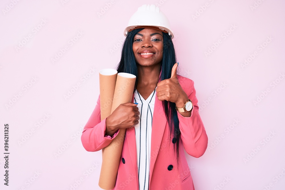 Young african american woman wearing architect hardhat holding blueprints smiling happy and positive, thumb up doing excellent and approval sign