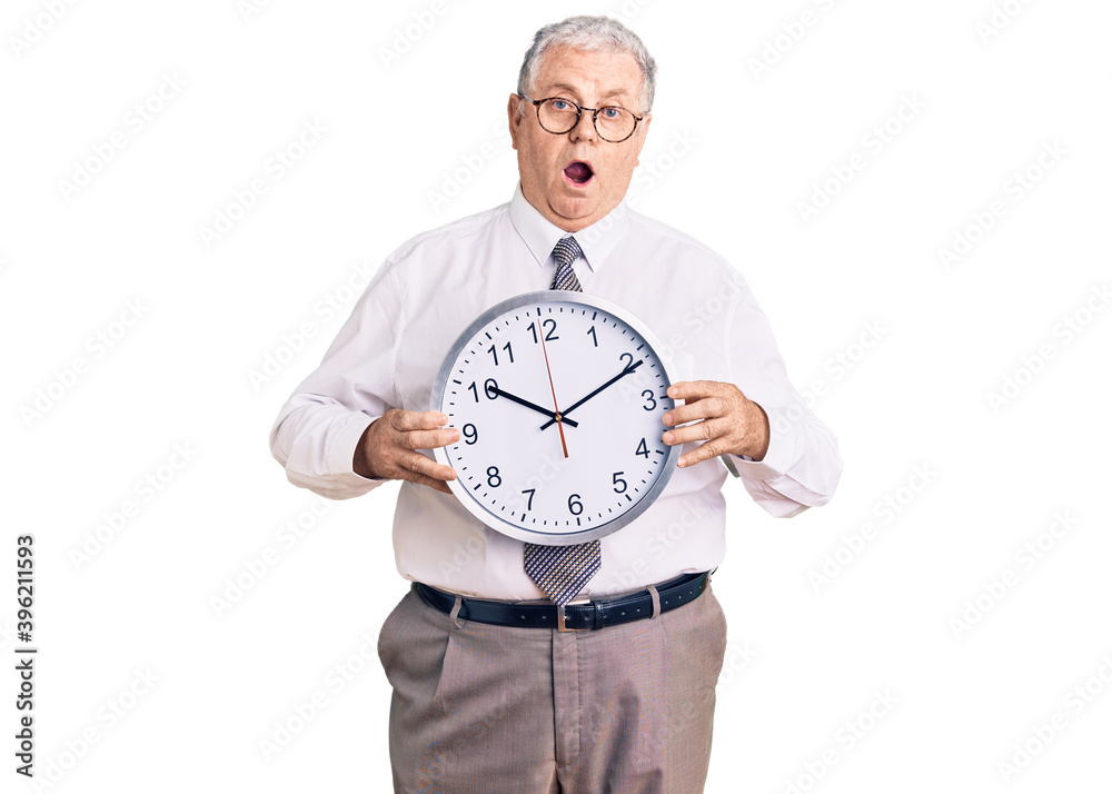 Senior grey-haired man wearing business clothes and holding clock scared and amazed with open mouth for surprise, disbelief face Senior grey-haired man wearing business clothes and holding clock scared and amazed with open mouth for surprise, disbelief face