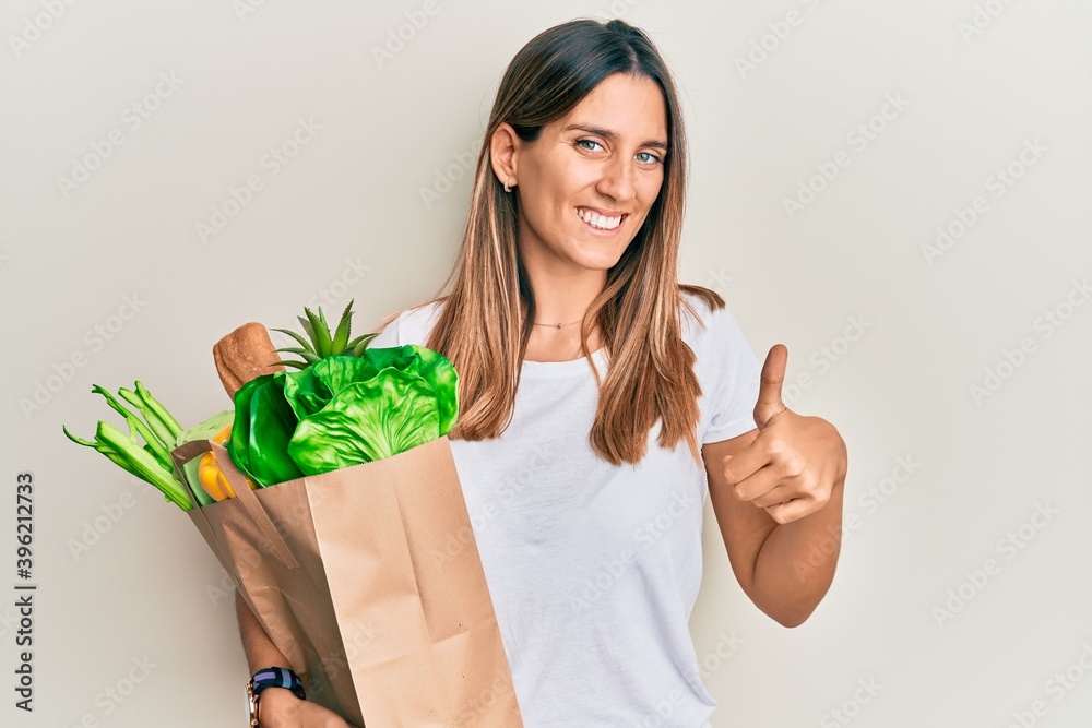 Brunette young woman holding paper bag with bread and groceries smiling happy and positive, thumb up doing excellent and approval sign