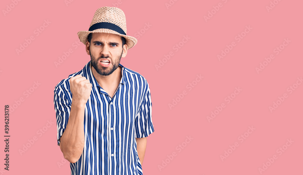 Young handsome man with beard wearing summer hat and striped shirt angry and mad raising fist frustrated and furious while shouting with anger. rage and aggressive concept. Young handsome man with beard wearing summer hat and striped shirt angry and mad raising fist frustrated and furious while shouting with anger. rage and aggressive concept.