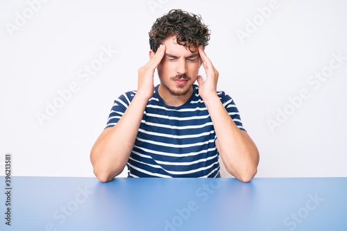 Young caucasian man with curly hair wearing casual clothes sitting on the table suffering from headache desperate and stressed because pain and migraine. hands on head.