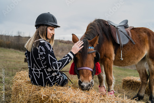 Young caucasian blonde woman female girl pet and play with the horse riding in nature wearing helmet in winter or autumn day against a cloudy sky in the field - Freedom friendship concept copy space © Miljan Živković