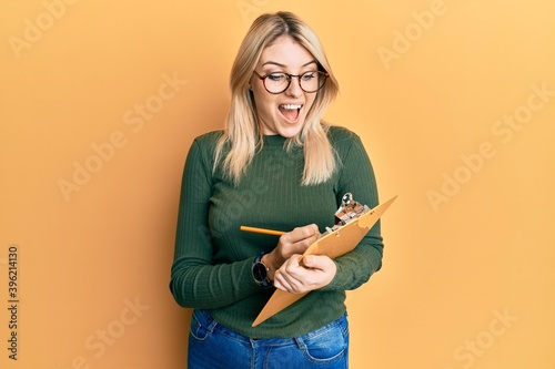 Young caucasian woman writing on checklist clipboard celebrating crazy and amazed for success with open eyes screaming excited.