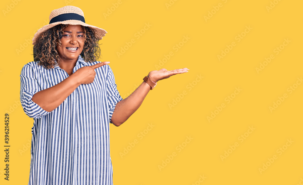 Young african american plus size woman wearing summer hat amazed and smiling to the camera while presenting with hand and pointing with finger.
