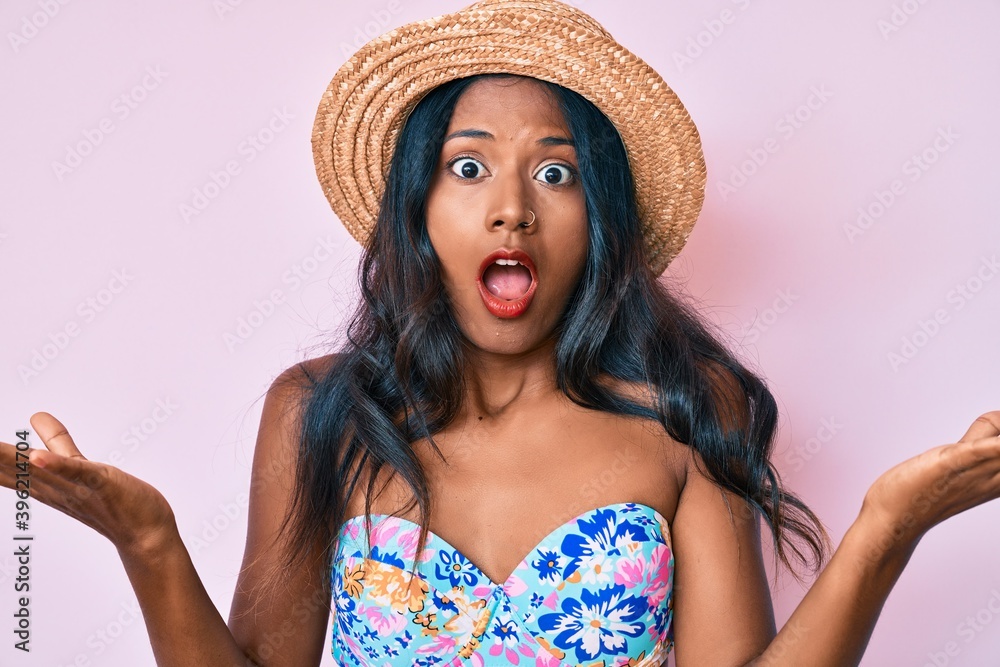 Young indian girl wearing bikini and summer hat clueless and confused expression with arms and hands raised. doubt concept. Young indian girl wearing bikini and summer hat clueless and confused expression with arms and hands raised. doubt concept.