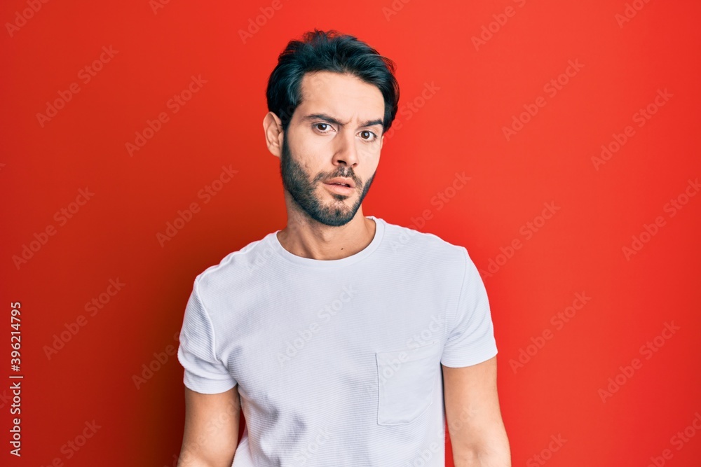 Young hispanic man wearing casual white tshirt in shock face, looking skeptical and sarcastic, surprised with open mouth Young hispanic man wearing casual white tshirt in shock face, looking skeptical and sarcastic, surprised with open mouth