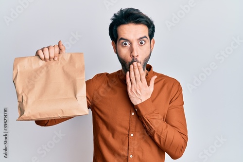 Young hispanic man holding take away food covering mouth with hand  shocked and afraid for mistake. surprised expression