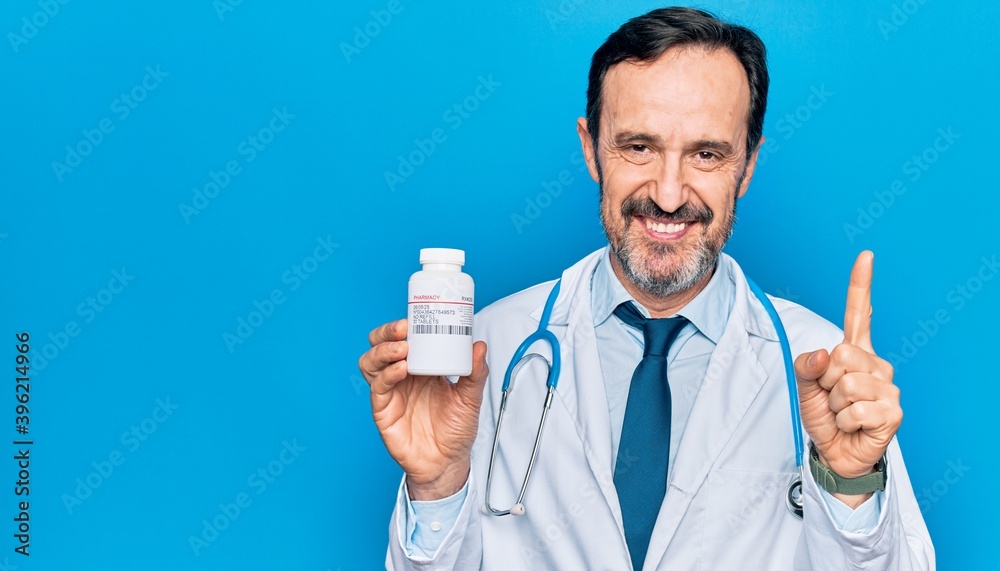 Middle age handsome doctor man wearing stethoscope holding bottle of pills smiling with an idea or question pointing finger with happy face, number one