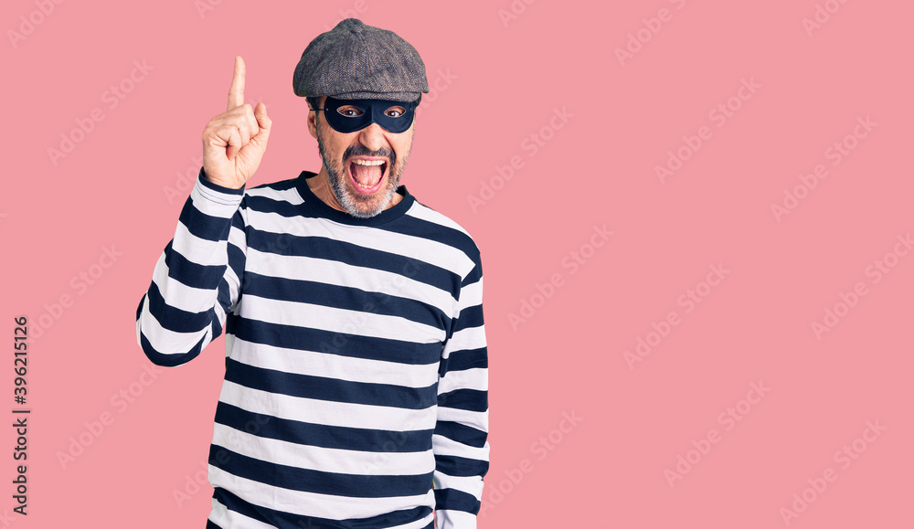 Middle age handsome man wearing burglar mask pointing finger up with successful idea. exited and happy. number one. Middle age handsome man wearing burglar mask pointing finger up with successful idea. exited and happy. number one.
