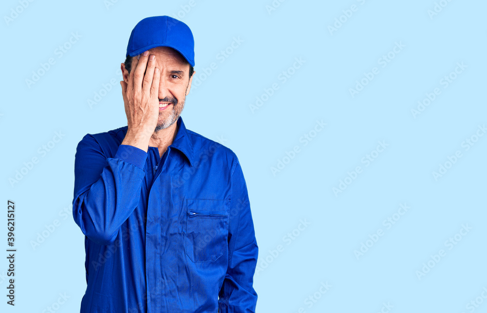 Middle age handsome man wearing mechanic uniform covering one eye with hand, confident smile on face and surprise emotion.