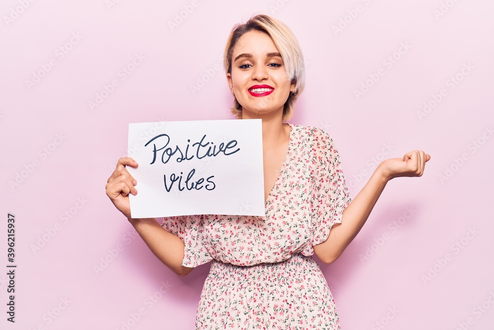 Young beautiful blonde woman holding positive vibes banner screaming proud, celebrating victory and success very excited with raised arm Young beautiful blonde woman holding positive vibes banner screaming proud, celebrating victory and success very excited with raised arm