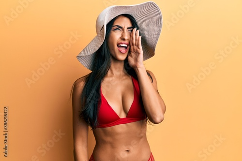 Beautiful hispanic woman wearing bikini and summer hat shouting and screaming loud to side with hand on mouth. communication concept.
