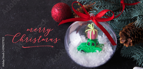Congratulation of merry christmas on dark background with transparent christmas ball with a toy car gift on the roof in the snow