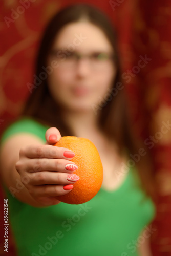 A girl with a beautiful orange manicure is holding a ripe orange in her hands. Blurred face in the background. Hand in the foreground.