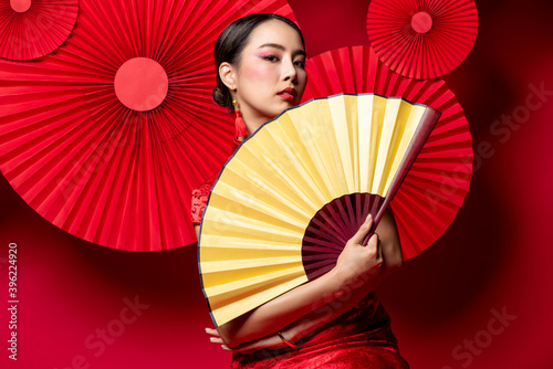 Beautiful Asian woman in traditional Chinese dress with colorful make up holding golden fan in oriental style red background