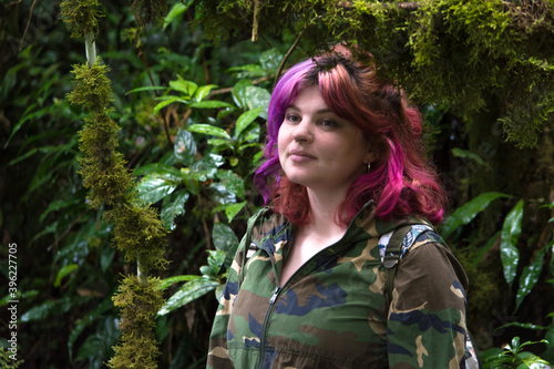 Young woman with pink hair and camouflage jacket, looking at camera in the middle of a green tropical rainforest, with plants and trunks branches with moss