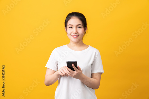 Cheerful beautiful Asian woman holding smartphone on light yellow background. © sitthiphong