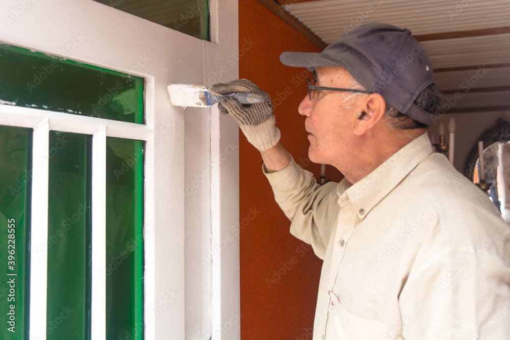 Old man with beige shirt and grey cap, holding a brush with white paint and painting a door with green windows