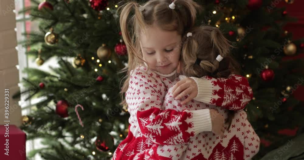 Two beautiful little sisters hugging with a Christmas tree in the background. Happy holiday concept Two beautiful little sisters hugging with a Christmas tree in the background. Happy holiday concept