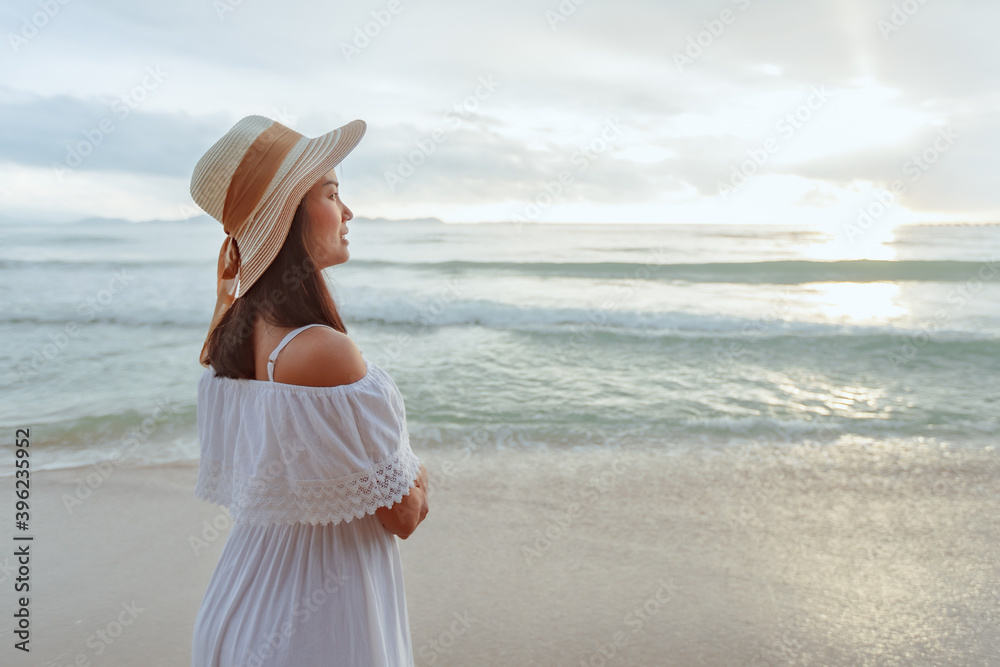 Asian woman wearing stylish hat and clothes looking to the ocean and enjoying beautiful Sunset on the Beach. Summer holidays and travel concept.