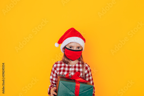 Child holding a box in green packaging with a gift. Christmas mood. She has a santa hat on her head and a medical mask on her face