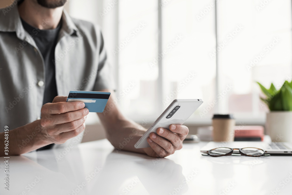 Fototapeta premium Man holding credit card and using smartphone at office, businessman shopping online, e-commerce, internet banking, spending money, working from home concept