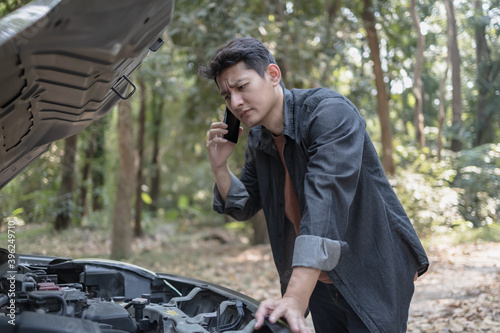 Man usea cellphone call garage in front of the open hood of a broken car on the road in the forest. Car breakdown concept.