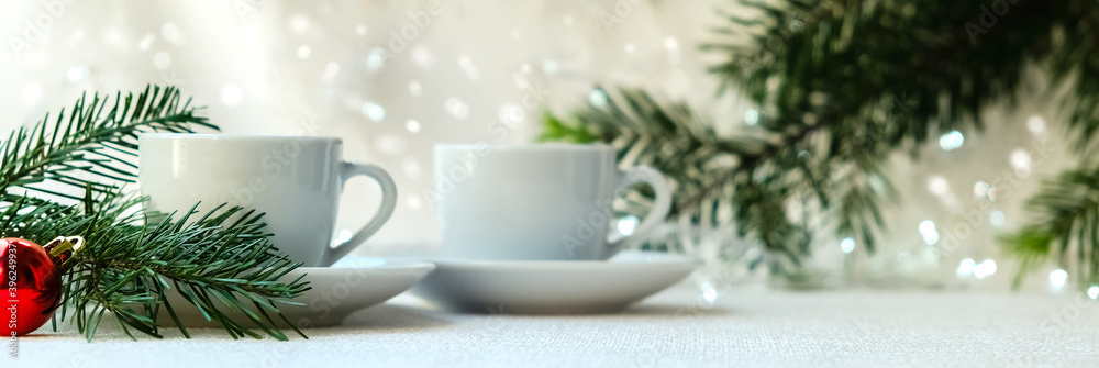 two white coffee cups on white background with garland. Near the branch of spruce and red ball. The concept of Christmas