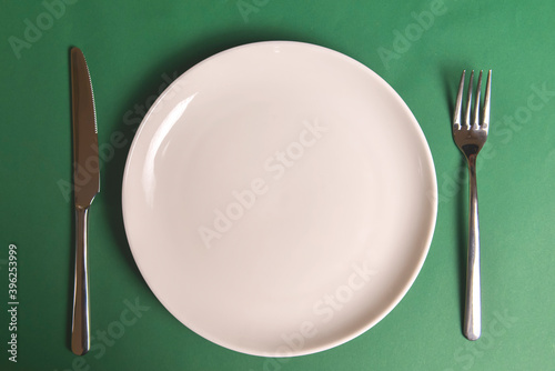 Vintage silverware on dark green background. White plate with knife and fork over green table, flat lay.