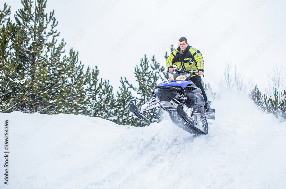Jumping on a snowmobile on a background of winter forest. Bright snowmobile. Athlete on a snowmobile moving in the winter forest in the mountains. Man and fast action snowmobile jumping