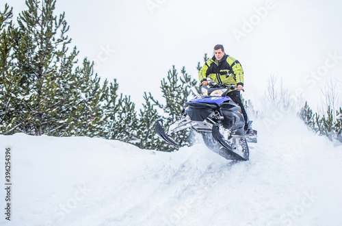 Jumping on a snowmobile on a background of winter forest. Bright snowmobile. Athlete on a snowmobile moving in the winter forest in the mountains. Man and fast action snowmobile jumping © Yevhen