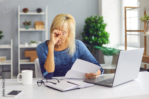 Upset senior woman sitting at desk, holding bankruptcy or debt notification from bank photo