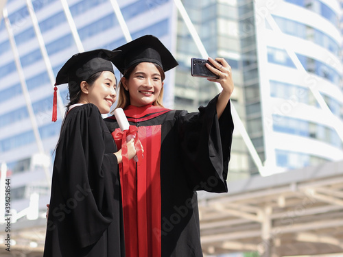 happy graduated women in graduation gowns making selfie photo with mobile phone. City building background. Education, Friendship and technology concept..