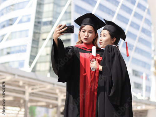 happy graduated women in graduation gowns making kissing lips, taking selfie photo with mobile phone. City building background. Education, Friendship and technology concept..