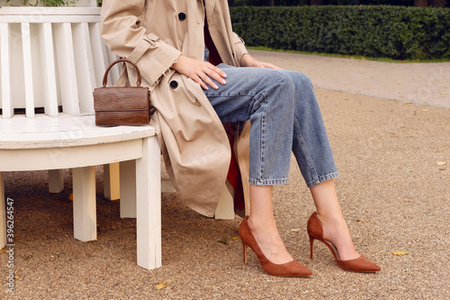 Closeup legs high heels, woman in beige coat and blue jeans with brown leather bag. Fashion