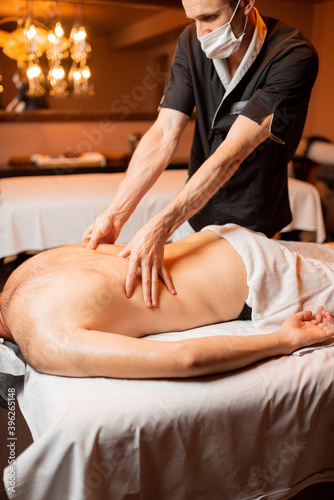 Professional masseur in facial mask doing a deep massage to a male client at Spa salon. Business during the epidemic concept