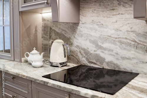 beautiful closeup of custom designed kitchen, with marble looking quartz countertop and backsplash. cream electric kettle with porcelain tea accessories on the marble countertop next to ceramic hob