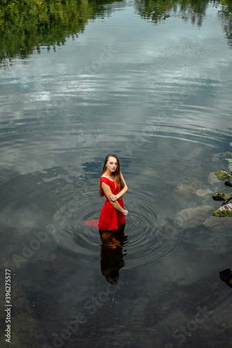 Vertical portrait of young pretty wet woman mermaid standing in the water river or lake touching clothes, dressed in red dress, looking at the camera, copy space and nature blur background. 