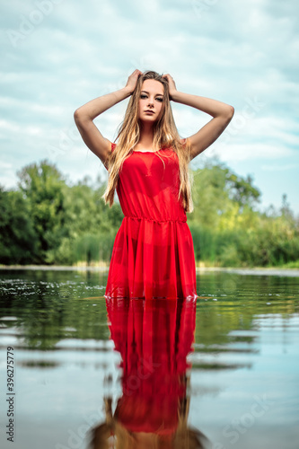 Vertical portrait of young pretty wet woman mermaid standing in the water river or lake touching head, dressed in red dress, looking at the camera, copy space and nature blur background. 