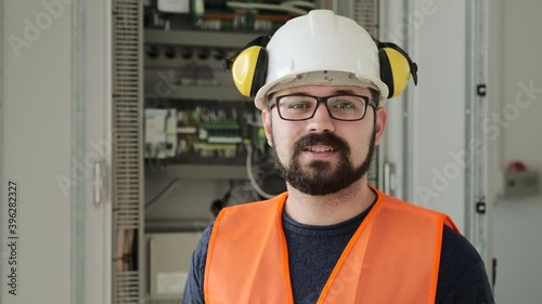 Bearded worker in white helmet with headphones stands near switchboard cabinet at electrical substation before start closeup