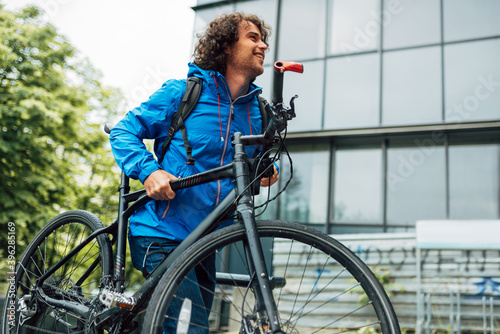 Side view outdoor image of a young man with curly hair climbing on the building's stairs with his bike in his hands. Male courier with curly hair delivers a parcel with a bicycle in the city.