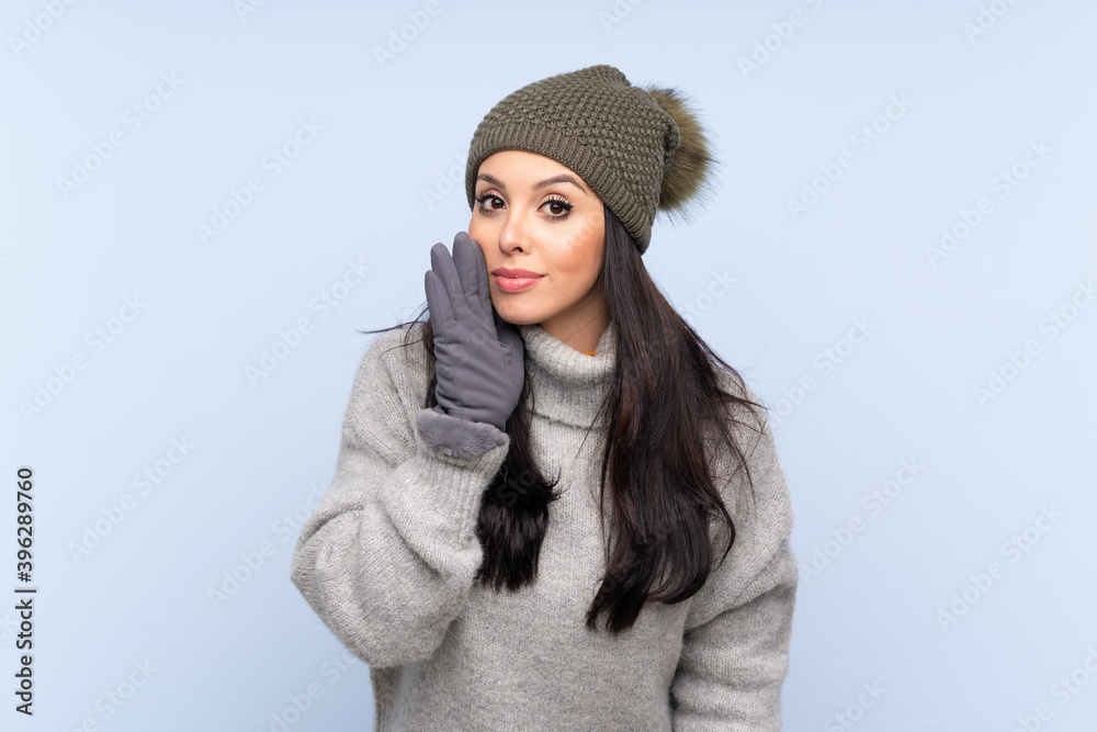 Young Colombian girl with winter hat over isolated blue background whispering something Young Colombian girl with winter hat over isolated blue background whispering something